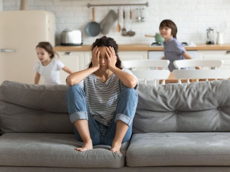 Maman épuisée assise sur un canapé avec ses deux enfants turbulents en arrière-plan, illustrant les défis du TDAH au quotidien.