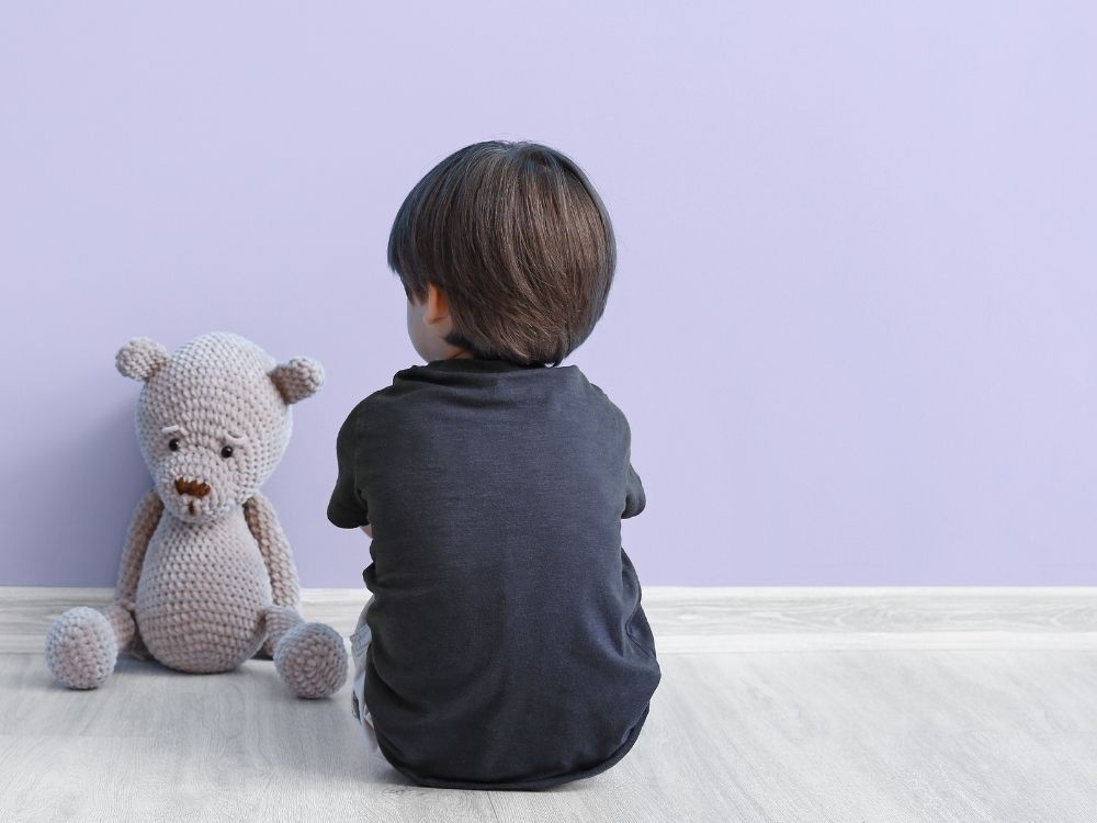 Jeune garçon assis calmement de dos avec son doudou face à un mur violet, illustrant la distractibilité et l'hypoactivité dans le TDAH.