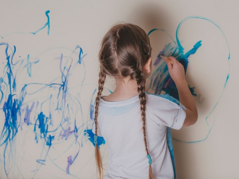 Jeune fille avec des tresses dessinant avec application sur un mur blanc, représentant l'impulsivité et le non-respect des règles chez un enfant TDAH.