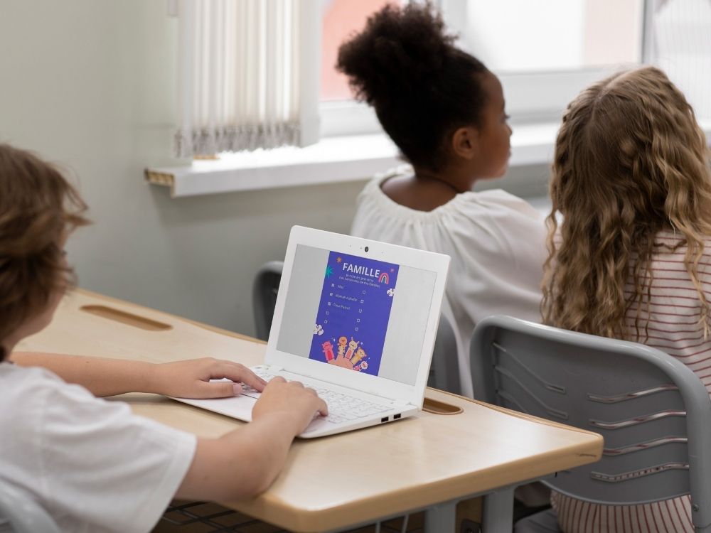 Trois enfants assis à des pupitres d'école, vus de dos. Au premier plan, un garçon tape sur un ordinateur portable blanc dont l'écran affiche une illustration violette intitulée "FAMILLE"