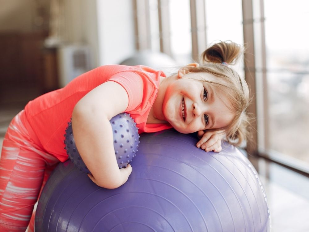 Petite fille souriante allongée sur un gros ballon de gym violet pendant un atelier pratique d'Harmonie Apprentissage.