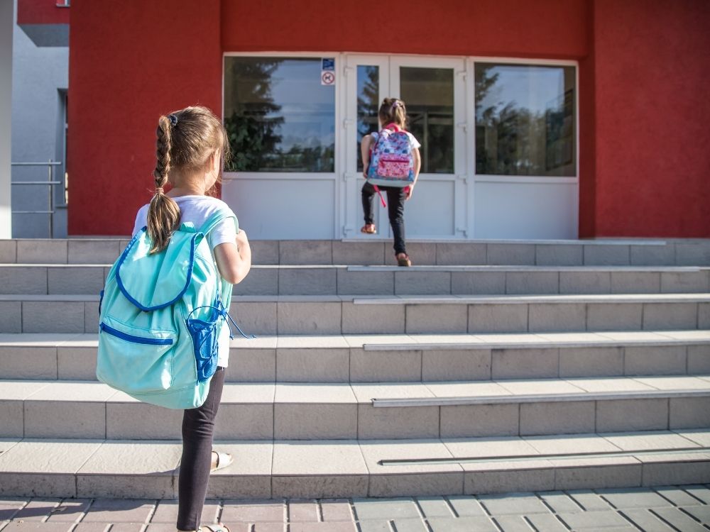 Deux jeunes enfants avec des cartables montant les marches de l'entrée de l'école, illustrant l'inclusion scolaire et l'accès à l'éducation.