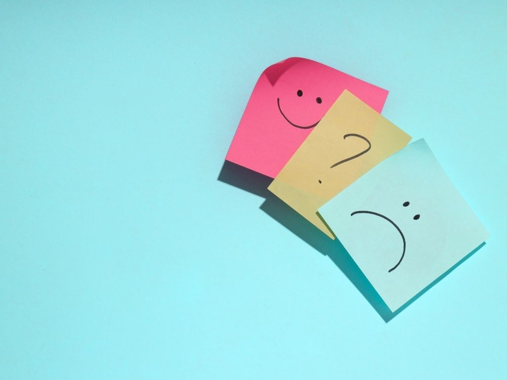 Vue de dessus de trois notes adhésives superposées sur un fond turquoise, illustrant le processus de régulation émotionnelle : une note rose avec un visage souriant (la joie), une note jaune avec un point d'interrogation (l'incertitude), et une note bleue avec un visage triste (la tristesse).