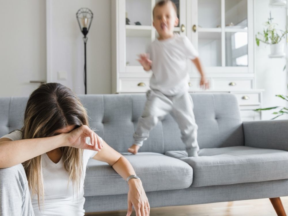 Maman épuisée sur un canapé, cachant son visage, pendant que son jeune enfant hyperactif saute derrière elle, illustrant les défis quotidiens du TDAH et l'utilité de la guidance parentale d'Harmonie Apprentissage.