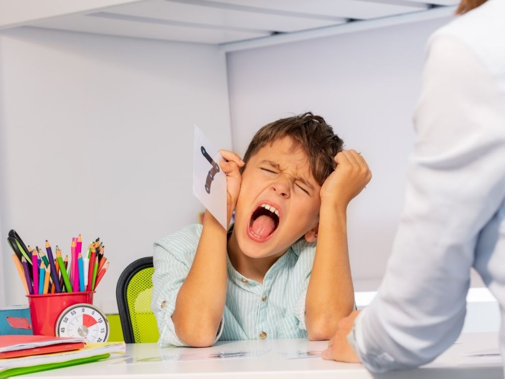 Jeune garçon fatigué ou frustré qui crie à son bureau face à une intervenante, illustrant les difficultés d'attention et de gestion des émotions.