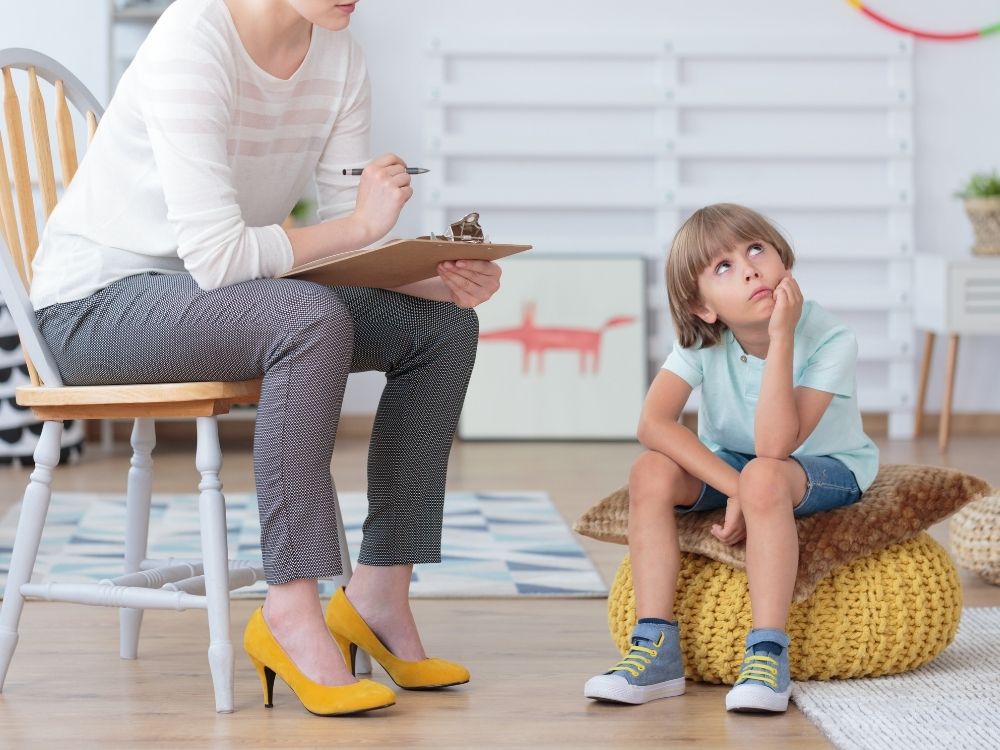 Jeune enfant assis sur un pouf en consultation avec une professionnelle qui prend des notes, illustrant l'accompagnement des troubles du neurodéveloppement.