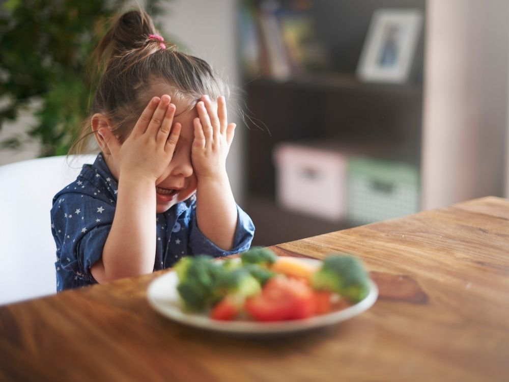 Enfant assise à table devant son assiette de légumes se cachant le visage dans les mains