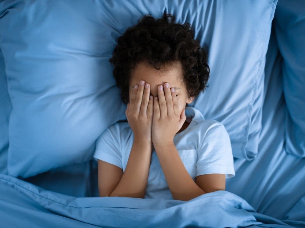 Enfant couché dans son lit se cachant le visage avec ses mains (Troubles du sommeil)