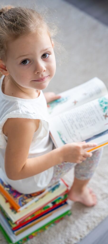 Petite fille assise par terre lisant un livre, à côté d'une pile de livres